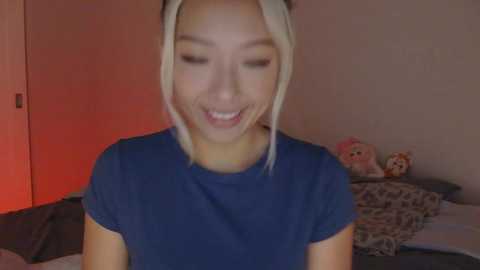 Video of a young Asian woman with straight, platinum blonde hair, wearing a blue t-shirt, smiling with closed eyes in a dimly lit room.