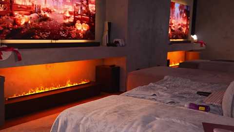 Video of a dimly lit bedroom with two large TVs displaying warm, red-lit holiday scenes. Beneath each TV, a glowing fireplace effect is visible. The bed features light bedding and a remote control on the pillow.