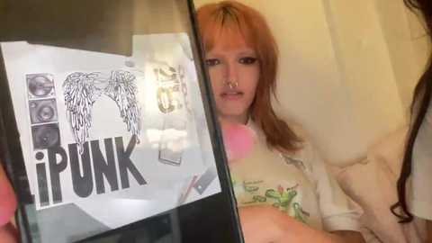 Video of a young woman with light skin and reddish-brown hair, wearing a white t-shirt with green and yellow graphics, standing in front of a mirror. The mirror reflects a black \"I PUNK\" poster with a winged skull design.
