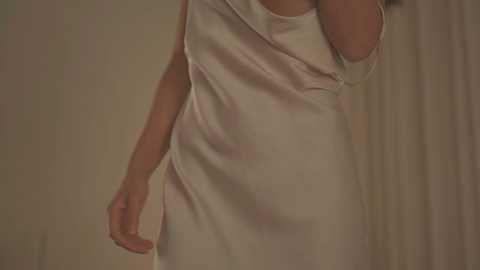 Video of a woman with light skin, wearing a white, off-shoulder satin dress, slightly lifting her arm. Background features beige curtains. The image captures the elegant, smooth texture of the fabric.