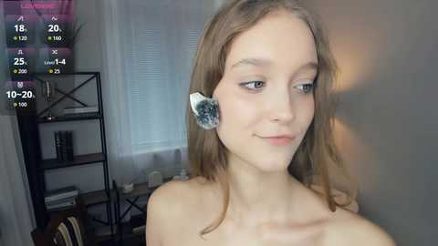 Video of a young, light-skinned woman with long, light brown hair, wearing a white, fluffy ear clip, standing topless in a modern, minimalist room with white walls and a black metal shelf.