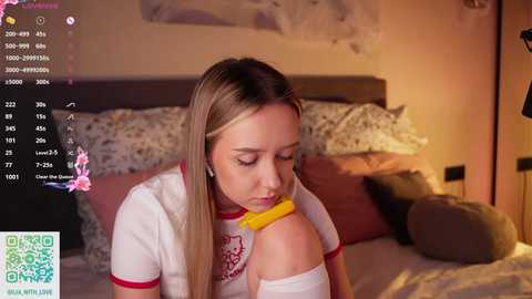Media: Video of a blonde woman with fair skin, wearing a white T-shirt with red trim, biting a yellow banana. Background shows a cozy bedroom with a bed, floral pillows, and soft lighting.