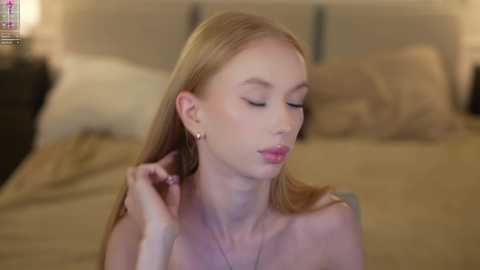 Media: Video of a young, slender, fair-skinned woman with long blonde hair, wearing small gold hoop earrings and pink lipstick, sitting on a bed in a dimly lit room.