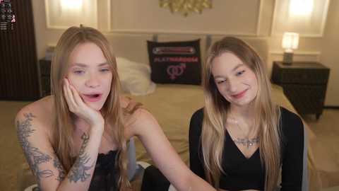 Media: Video of two young, fair-skinned women with long blonde hair, one in a black lace dress, the other in a black top, sitting on a beige bed with floral pillows.