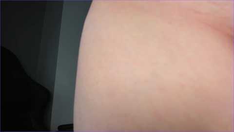 A close-up video of a pale, smooth human buttock, with a hint of a dark shadow in the background, possibly indicating a dark-colored object or person. The skin appears slightly textured with natural imperfections.