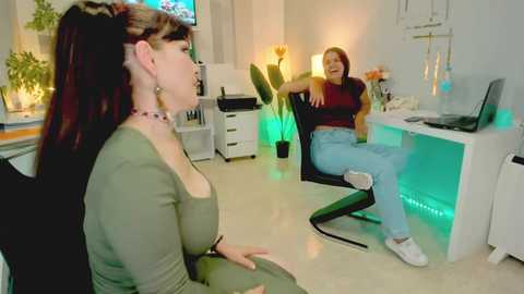 Video of a woman in a green top and jeans watching a woman in a red shirt and jeans on a glowing desk chair in a brightly lit, modern office.