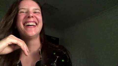 A candid video captures a smiling woman with long brown hair, wearing a black shirt, in a dimly lit room with textured walls.