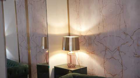 Video of a chic, modern living room with a marble-patterned wallpaper, a metallic lamp with a pink base, and a dark green tufted sofa.