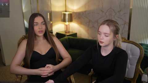 Video of two young women, one with long brown hair in a black strapless dress, the other with blonde hair in a black long-sleeve top, seated on a green velvet couch with a gold-framed mirror and a lit lamp behind them.