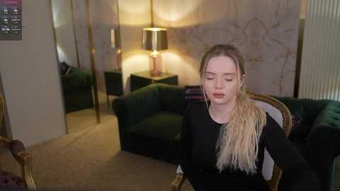 Video of a young, light-skinned woman with long blonde hair, wearing a black dress with a white feathered shoulder, standing in a dimly lit, retro-themed living room with green leather couches, gold lamps, and floral wallpaper.