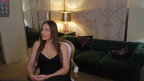 Video of a woman with long brown hair, fair skin, and a slender physique, sitting in a green velvet armchair in a dimly lit living room. She wears a black dress, and the room features a gold-patterned wall, a dark green sofa, and a table lamp.