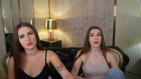 Video of two young women with long hair, one in a black spaghetti strap top, the other in a beige tank top, sitting on a dark green couch with a lamp and striped wallpaper in the background.