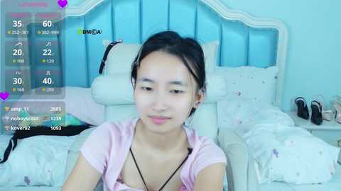 Video of an Asian woman with straight black hair, pink top, and black necklace, sitting on a white bed with a blue headboard. The room is softly lit with pastel decor.