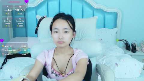 Video of an East Asian woman with straight black hair, wearing a pink top and black necklace, sitting on a bed with white linens and a blue upholstered headboard.