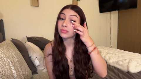 Video of a young woman with long brown hair, fair skin, and a slight pout, sitting on a bed with white and grey bedding, wearing a pink top.