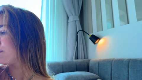 Media: Video of a young woman with straight brown hair, wearing a beige top, seated on a gray sofa. A black desk lamp with a warm light illuminates the room, featuring light blue curtains and white walls.