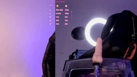 Video of a person wearing a black latex bodysuit, posing with a realistic silicone penis mounted on a black stand, illuminated by a ring light in a dimly lit studio.