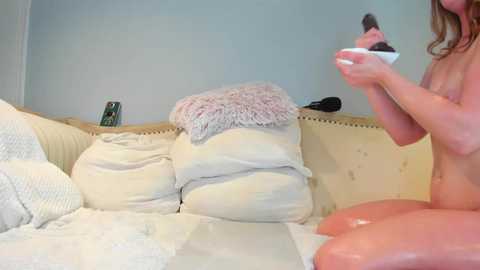 Video of a nude, fair-skinned woman with medium-length brown hair, holding a bowl of ice cream, sitting on a beige couch with cream blankets.