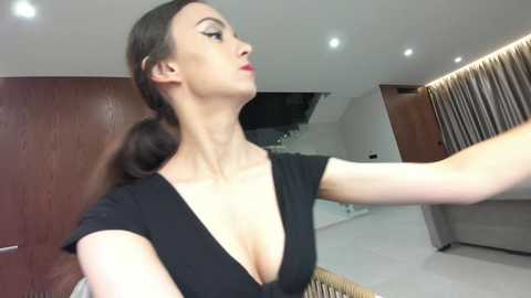 Video of a fair-skinned woman with long brown hair, wearing a black, deep V-neck dress, adjusting a radiator in a modern, minimalist room with wooden doors and white walls.