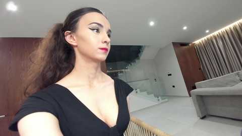 Video of a young woman with fair skin, long brown hair, and full lips, wearing a black dress. She stands in a modern, minimalistic living room with white walls, wooden furniture, and a grey couch.