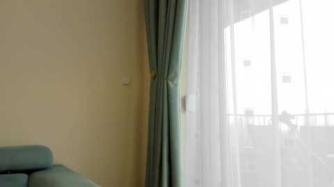 Video of a minimalist hotel room with a cream-colored wall, teal curtains, a light blue sofa, and a glass door leading to a balcony with a view of the city.