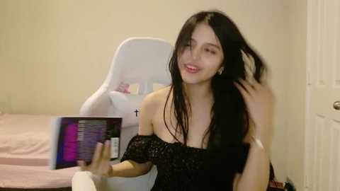 A video shows a young woman with long black hair, fair skin, and a petite frame, sitting in a white chair, smiling while holding a book in a simple, beige room.