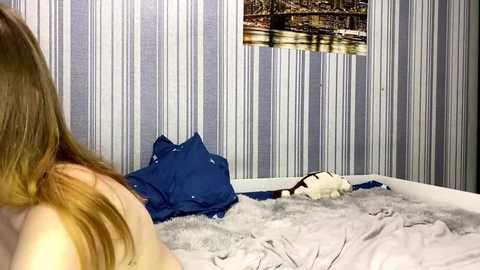 A video shows a woman with long, light brown hair lying on a bed with blue and white striped wallpaper. She is partially covered by a white blanket. A framed picture of a bridge is mounted on the wall.