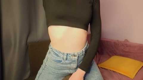 Video of a fair-skinned person in a black long-sleeve crop top, high-waisted light blue jeans, sitting on a pink sofa with a mustard-yellow cushion. Background includes a gray curtain and a pink wall.