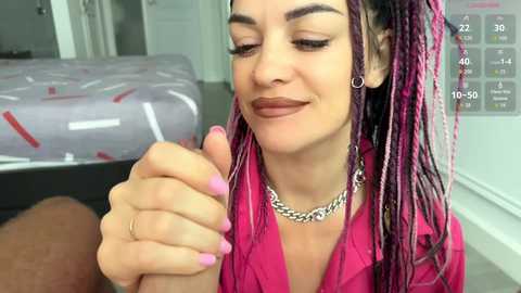 Video of a smiling woman with purple dreadlocks, pink nails, and silver jewelry, holding hands with a man's arm. She wears a pink shirt.