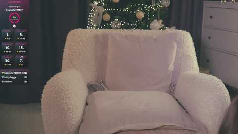 Video of a cozy, plush armchair covered in a soft, white, textured blanket. A Christmas tree with twinkling lights and ornaments decorates the background. The room is warmly lit, exuding a festive, intimate atmosphere.