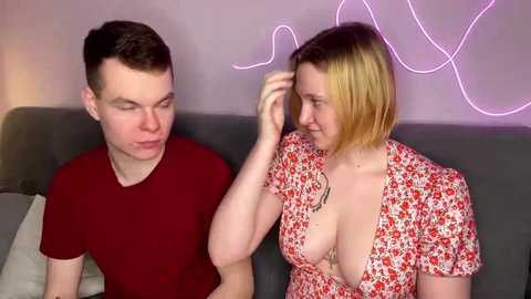 Video of a young couple on a grey couch. The man, with short brown hair, wears a red shirt; the woman, blonde with a short bob, has a low-cut floral dress, revealing cleavage. They smile at each other. Neon lights and a wall with abstract art are in the background.