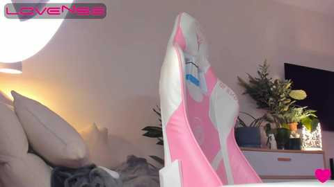 A video of a pink and white gaming chair in a cozy bedroom, featuring a large, round lamp, a TV, and a potted plant.