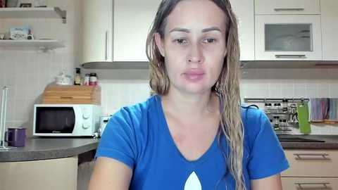 Video of a woman with long, blonde hair, wearing a blue t-shirt, standing in a modern kitchen with white cabinets and stainless steel appliances.