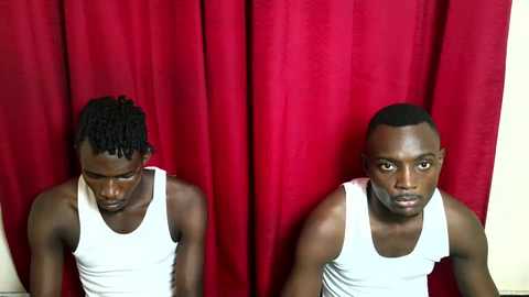 Video of two young Black men with medium skin tones, short hair, wearing white tank tops, standing against a deep red curtain backdrop, both looking downcast.