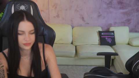 Media: Video of a young woman with long black hair, wearing a black tank top, seated in a black gaming chair in a modern living room with a white sectional sofa and purple wall.