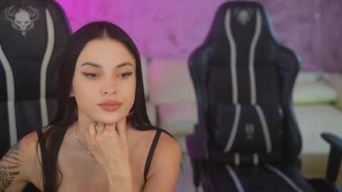 Video of a young woman with long dark hair, light skin, and a tattooed arm, seated in a gaming chair, wearing a black top, in a dimly lit room with purple lighting.