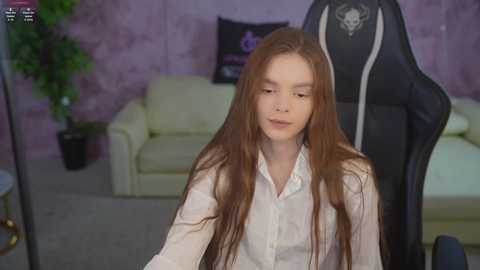Video of a young, slender, fair-skinned woman with long, straight brown hair, wearing a white button-up shirt, sitting on a black gaming chair in a cozy, pastel-pink room with a white sofa and green plant.