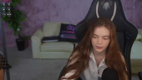 Video of a fair-skinned, long-haired woman with closed eyes, seated in a black gaming chair. Purple walls, white sofa, and a potted plant in the background.