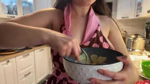 Video of an Asian woman with long black hair, wearing a pink and brown polka-dotted apron, mixing ingredients in a white bowl with a black handle in a modern kitchen.