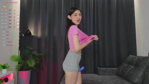Video of an Asian woman with long black hair, wearing a pink crop top and gray shorts, lifting her shirt, in a modern living room with a calendar, plants, and dark curtains.