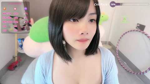 Video of a young Asian woman with straight black hair and light skin, wearing a blue cardigan, in a room with green and white plush toys.