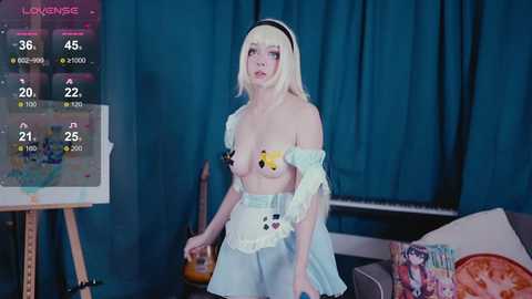 Video of a pale, slender woman with blonde hair and a white headband, wearing a revealing, off-shoulder Alice in Wonderland-themed dress with large pastel flowers covering her breasts. Background includes a blue curtain, a painting, and a gaming monitor displaying a virtual world.