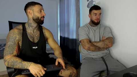 Video of a muscular, tattooed man in a black tank top, seated at a desk in a dimly lit room, with a confused-looking man in a gray t-shirt and sweatpants standing beside him.