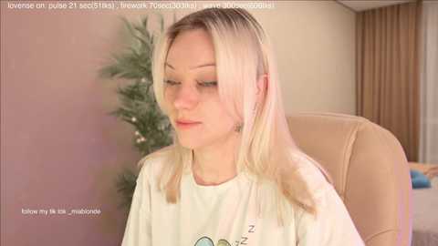 A video of a fair-skinned, blonde-haired woman with closed eyes, wearing a white T-shirt, seated on a beige chair in a softly lit room.