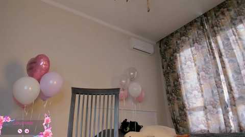 Video of a minimalist, beige-walled bedroom with a floral-patterned curtain, a white air conditioner, and a bed with a wooden headboard, featuring a cluster of pink and white heart-shaped balloons.
