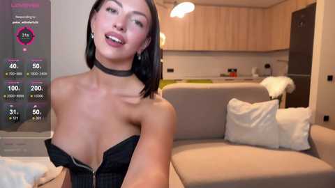 A video of a smiling, fair-skinned woman with shoulder-length black hair, wearing a black choker and unzipped black dress, standing in a modern living room with white couches.