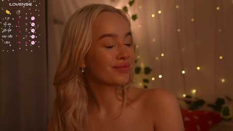 Video of a smiling, topless blonde woman with wavy hair, wearing a necklace, against a cozy, dimly lit room with string lights and a bed.