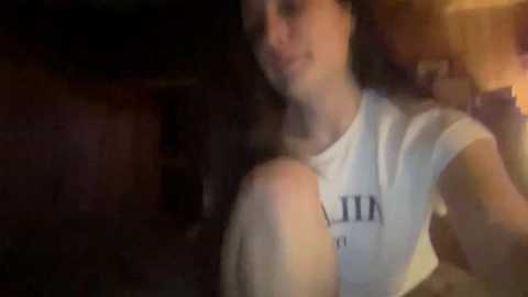 A blurry video of a young woman with curly dark hair, wearing a white t-shirt with text, standing in a dimly lit room with warm lighting.
