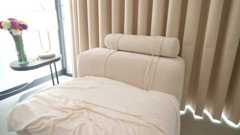 Video of a modern bedroom with a beige upholstered headboard, cream-colored bedding, and a vase of colorful flowers on a black side table near floor-to-ceiling beige curtains.