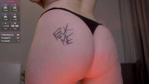 A close-up video of a light-skinned person's bare buttocks, wearing a black thong. A tattoo of a heart and arrow is visible on the left cheek. Background shows a bedroom with a TV screen displaying social media notifications.